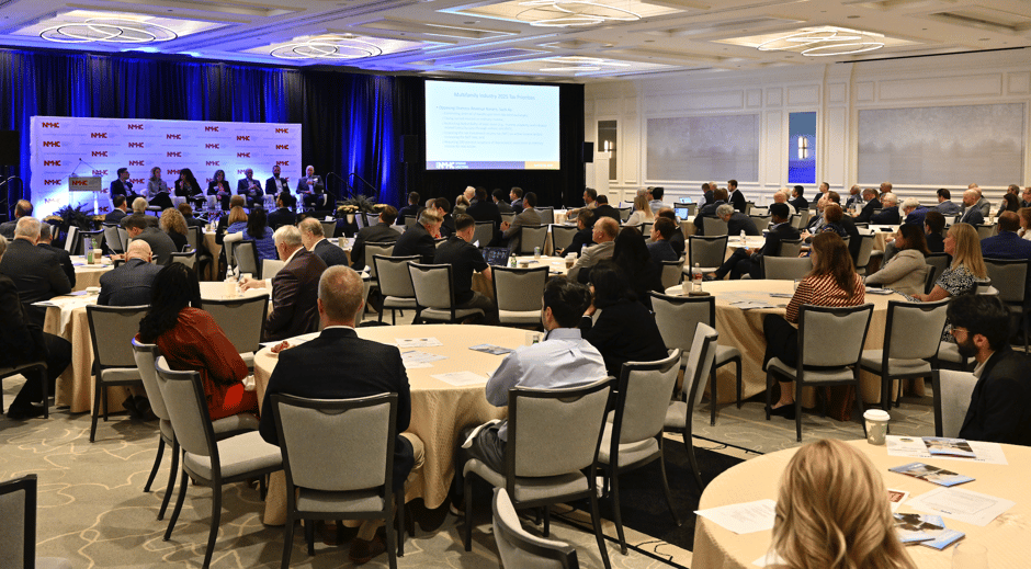 Attendees at the 2025 NMHC Spring Meeting sit in roundtable discussions during a panel on multifamily industry tax priorities, with a stage and large projection screen in view.